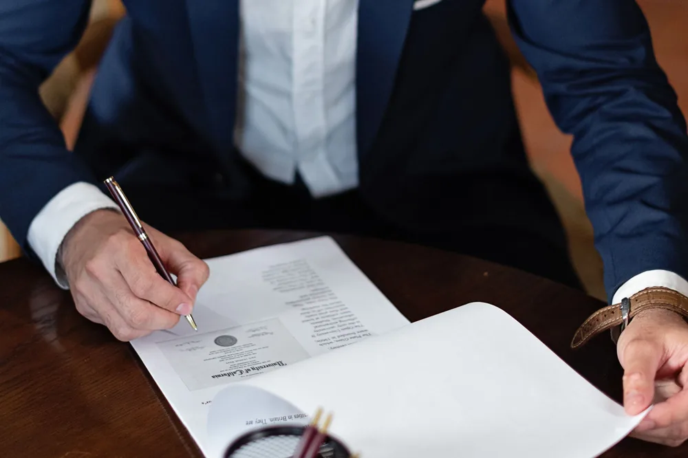 Homem de terno assinando um documento oficial com selo sobre a mesa