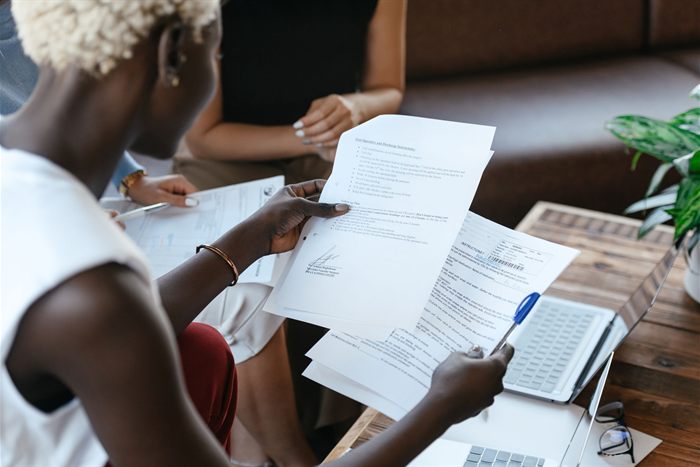 Duas pessoas sentadas à mesa examinando documentos impressos, uma delas segurando as páginas para leitura ao lado de um notebook