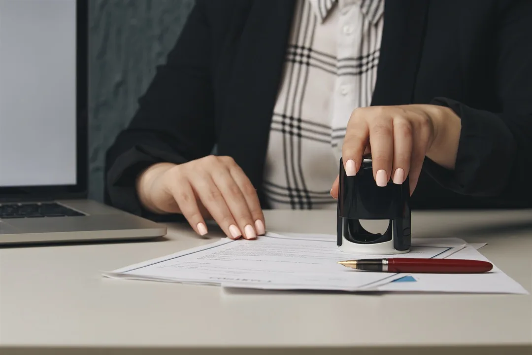 Mão feminina carimbando um documento sobre a mesa, representando a apostila e a autenticação de papéis oficiais
