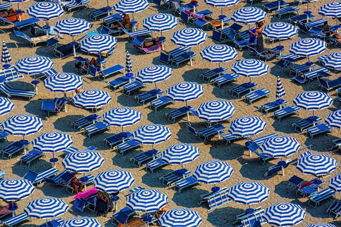 Vista aérea de dezenas de guarda-sóis listrados em azul e branco organizados em fileiras numa praia italiana lotada