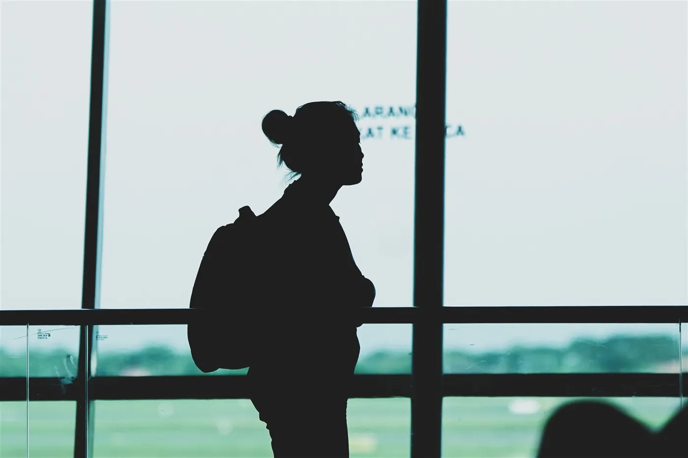 Silhueta de uma pessoa com mochila observando a pista de um aeroporto pela janela, momentos antes do embarque para o intercâmbio