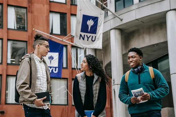 Três estudantes conversando em frente a um prédio universitário com bandeiras da NYU, em Nova York
