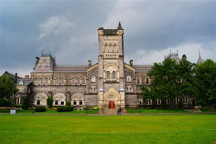 Fachada histórica em estilo gótico de um prédio universitário, com gramado verde em primeiro plano e céu nublado ao fundo