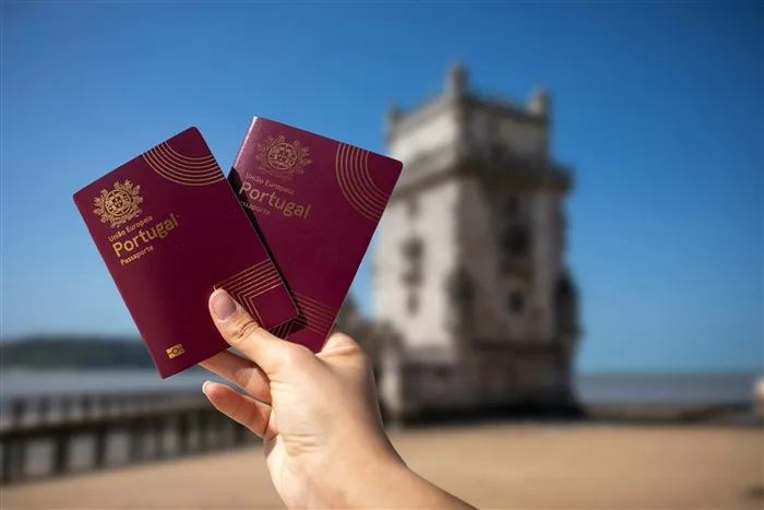 Mão segurando dois passaportes portugueses em frente à Torre de Belém, em Lisboa