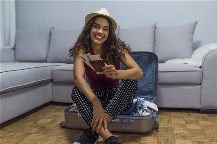 Mulher sorridente arrumando a mala com passaporte e passagens em mãos, sentada no chão de casa