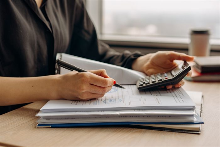 Mulher com uma calculadora na mão fazendo contas sobre uma pilha de documentos com selo oficial em uma mesa de escritório