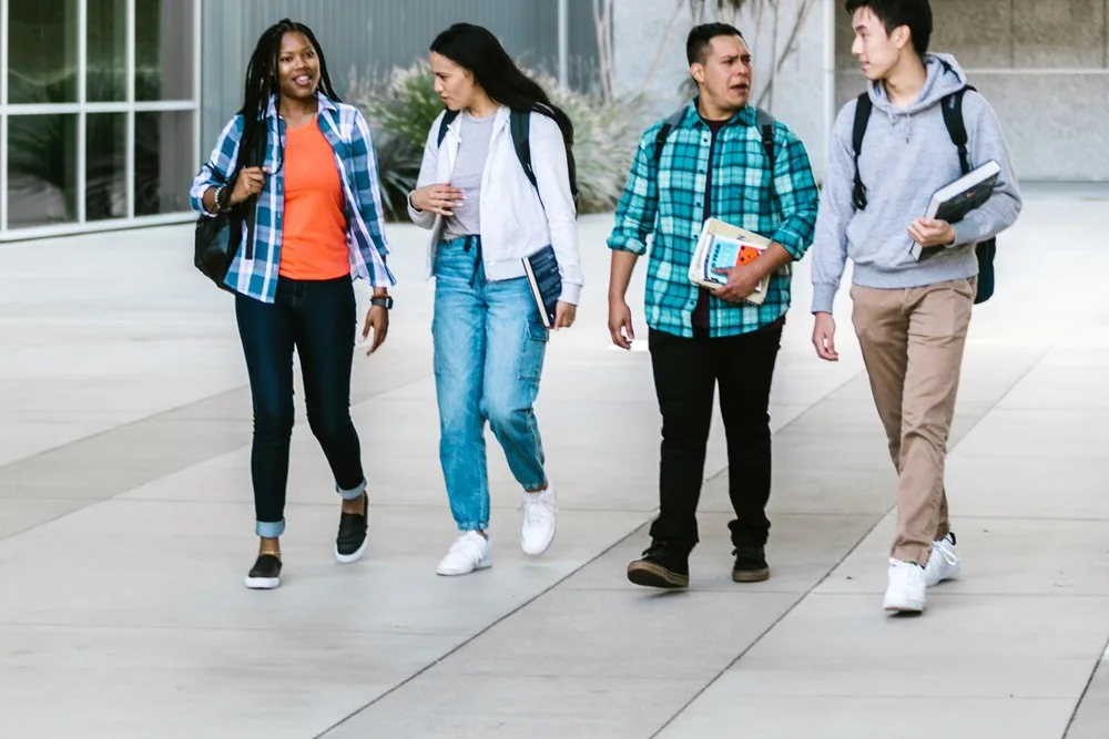 Quatro estudantes universitários conversando enquanto caminham por um pátio, com mochilas e livros nas mãos