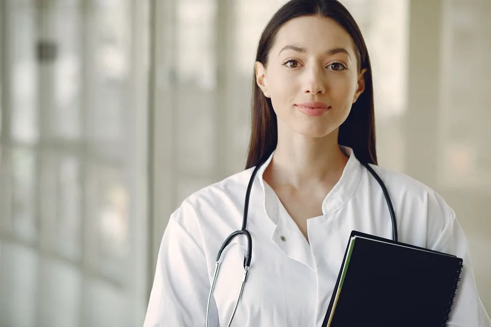 Médica de jaleco branco e estetoscópio, com um caderno sob o braço, olhando para a câmera num corredor claro