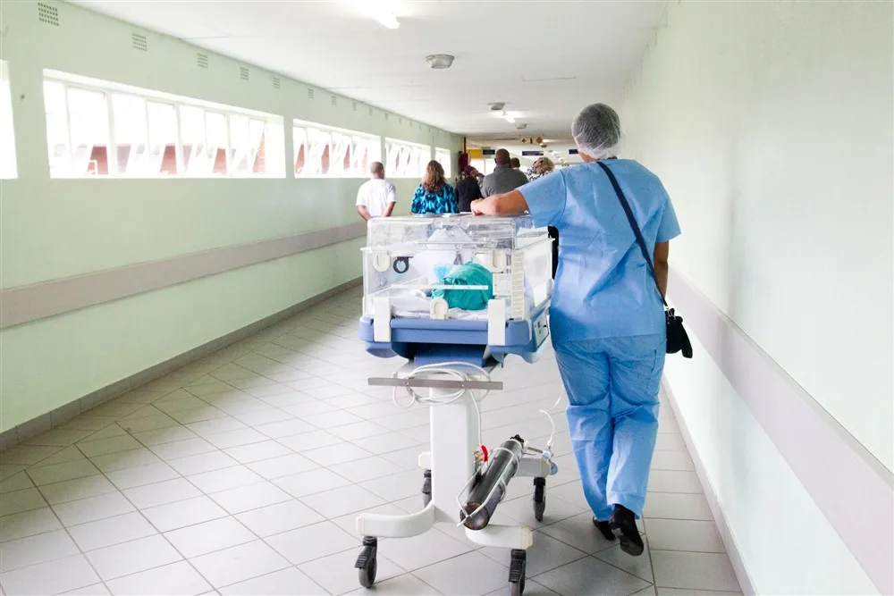 Profissional de saúde de uniforme azul empurrando uma incubadora por um corredor de hospital