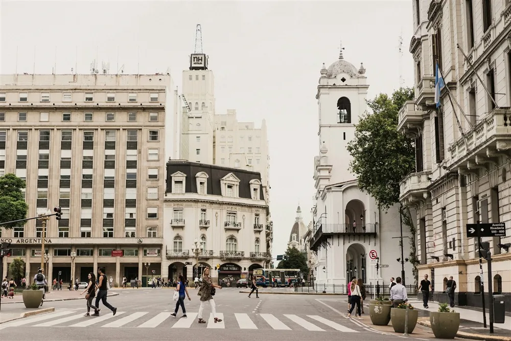 Pessoas atravessando uma rua em Buenos Aires, com prédios históricos e a bandeira da Argentina ao fundo
