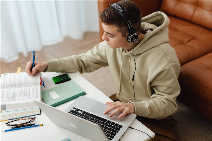 Jovem com fones de ouvido estudando em um notebook, cercado de livro, calculadora e óculos sobre a mesa