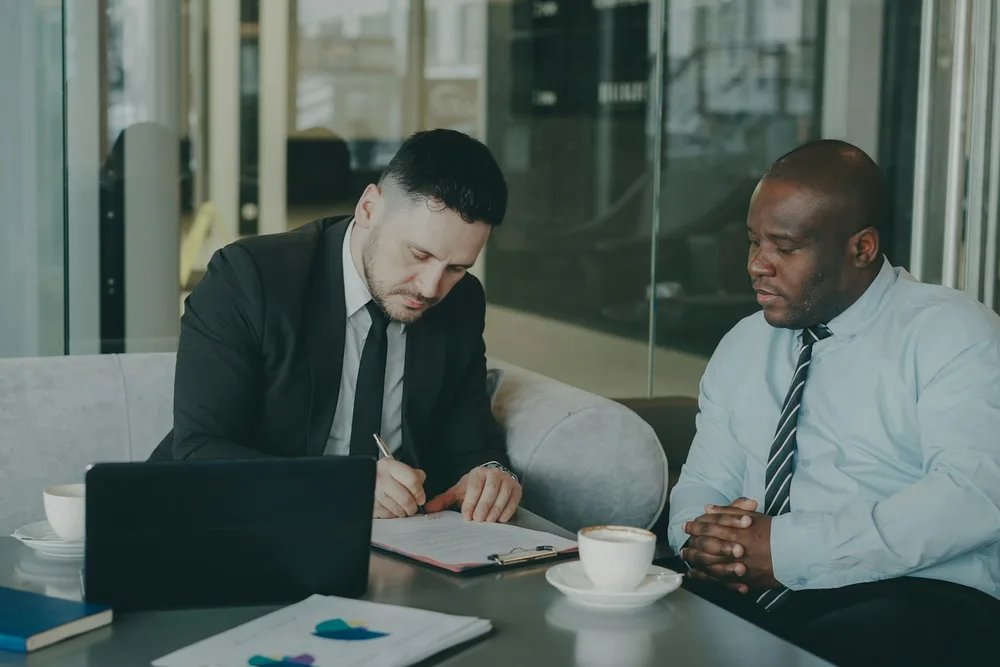 Dois executivos à mesa revisando um contrato, um deles assinando o documento ao lado de um notebook