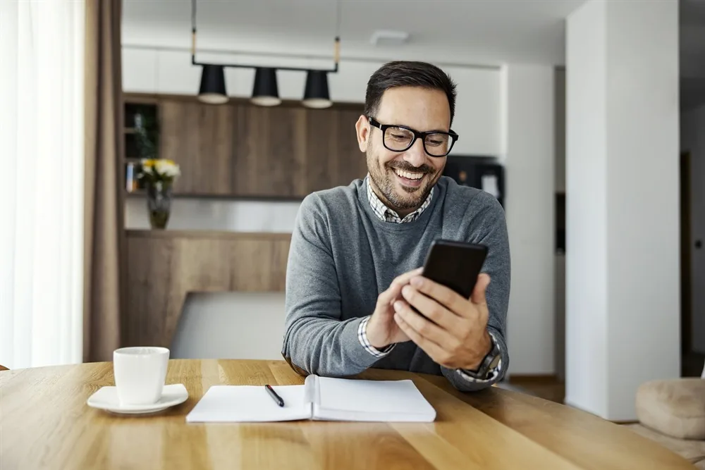 Homem sorrindo olhando o celular, sentado a uma mesa de madeira com caderno e xícara de café